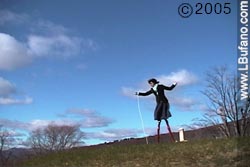 Lisa Bufano standng on tall stilts on a grassy hill, against a deep blue sky with fluffy clouds